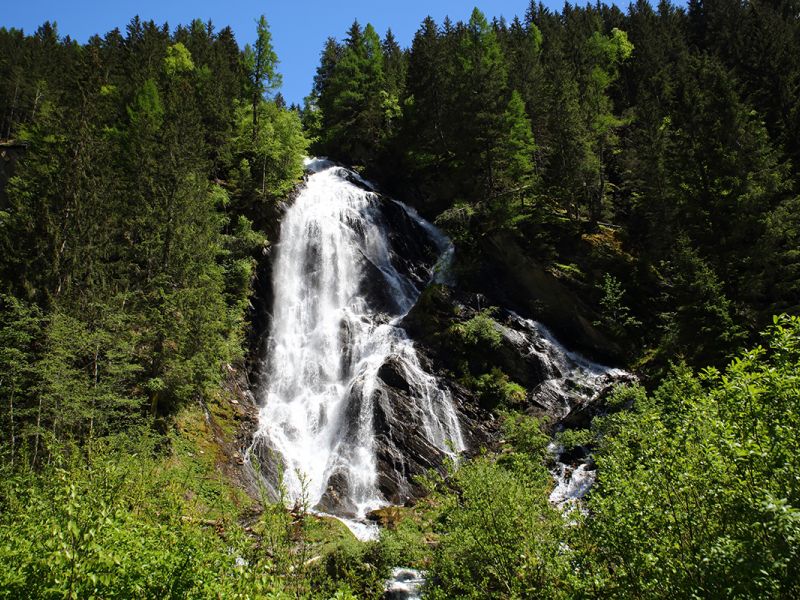 Ein kaskadenförmiger Wasserfall fließt einen felsigen Abhang hinunter, umgeben von üppig grünen Bäumen unter einem klaren blauen Himmel.