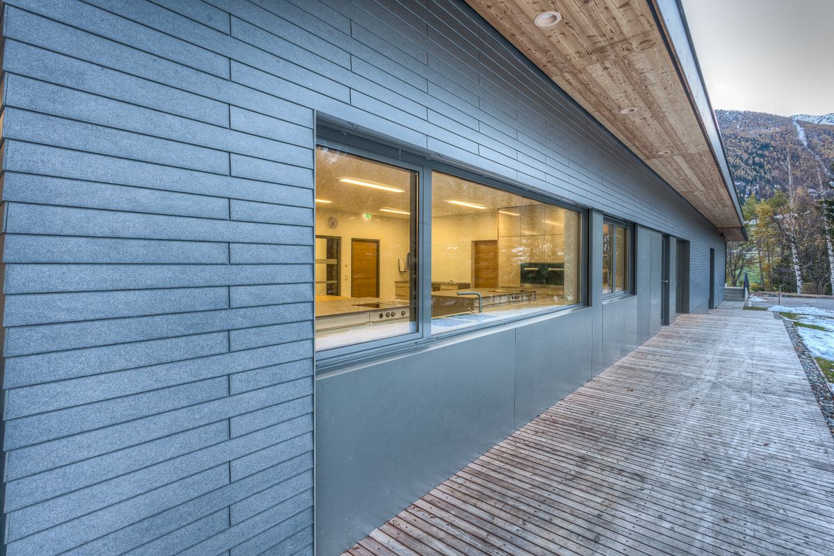 Modernes Gebäude mit Holzterrasse, großen Fenstern und schlichter grauer Verkleidung, mit den Bergen im Hintergrund.