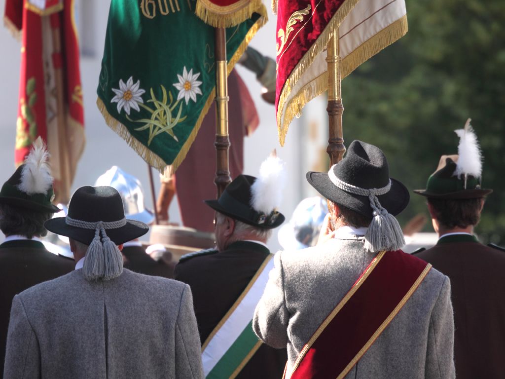 Eine Gruppe von Männern in traditioneller Kleidung, geschmückt mit Hüten und Schärpen, steht zusammen und hält bunte Fahnen während einer Parade.