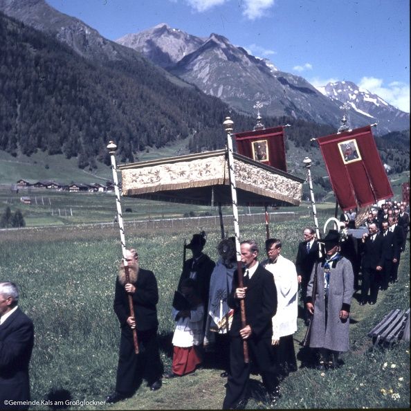 Eine Prozession von Menschen in formeller Kleidung, die in einer malerischen Berglandschaft spazieren gehen und kunstvolle Banner tragen.