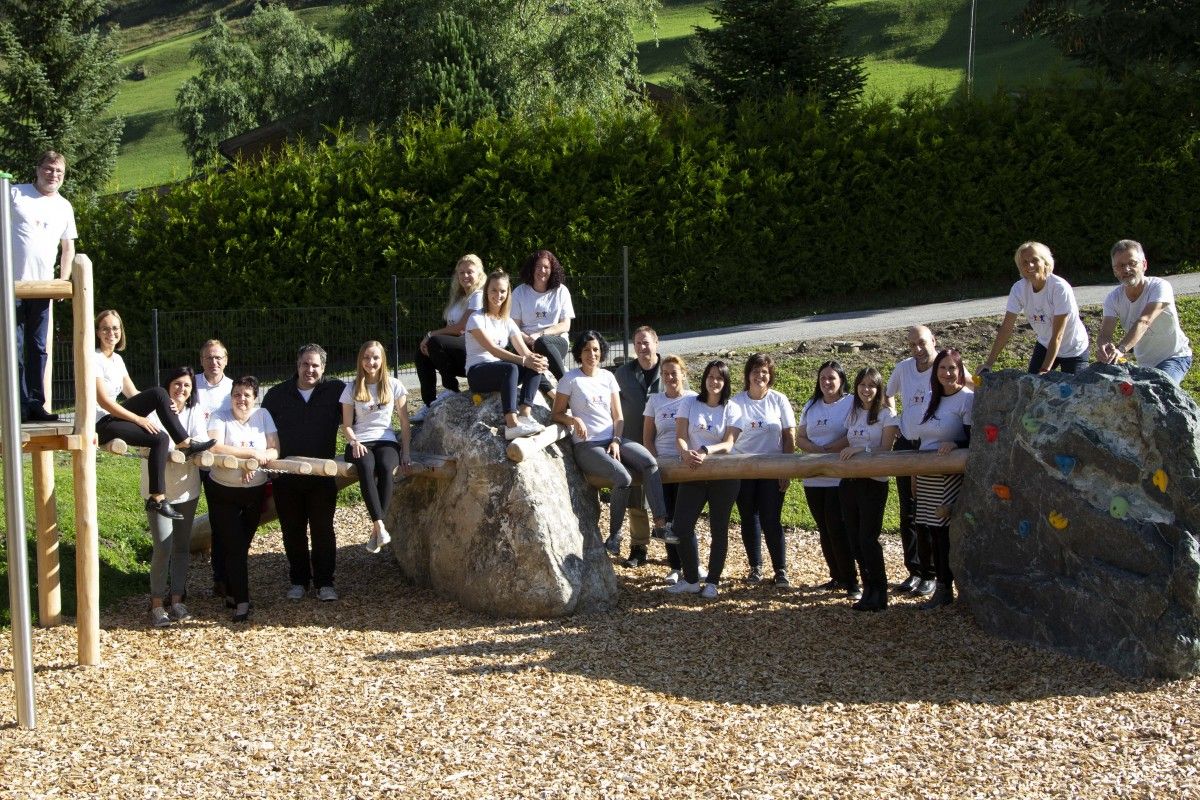 Eine Gruppe von Personen in passenden T-Shirts versammelt sich auf einem Spielplatz und posiert auf Felsen und Holzstrukturen in einer grünen Umgebung.
