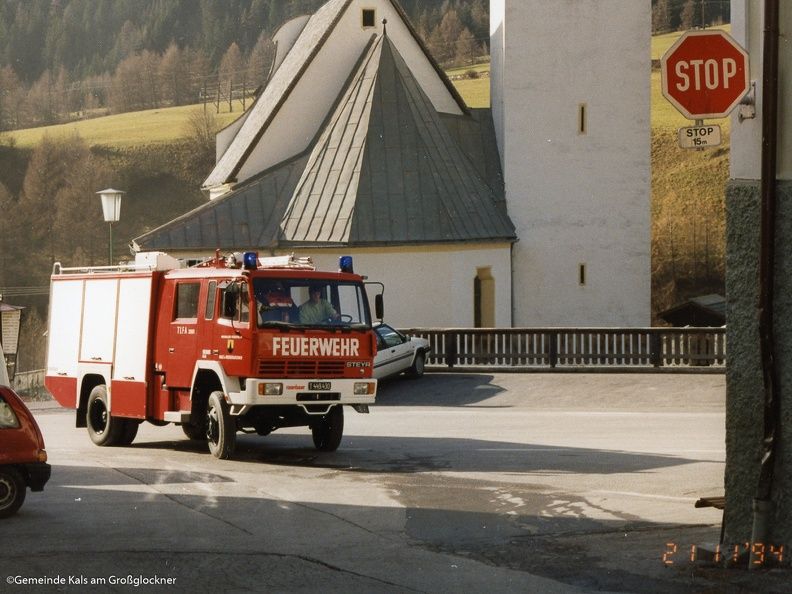 Ein altes rotes Feuerwehrauto mit der Aufschrift „FEUERWEHR“ nähert sich einer Kirche und einem Stoppschild in einem malerischen Bergdorf.