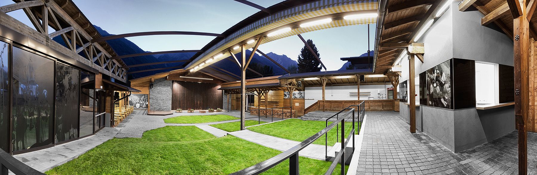Ein Panoramablick auf einen modernen Innenhof in Holzbauweise mit Grünflächen, Steinmauern und Bergen im Hintergrund in der Abenddämmerung.