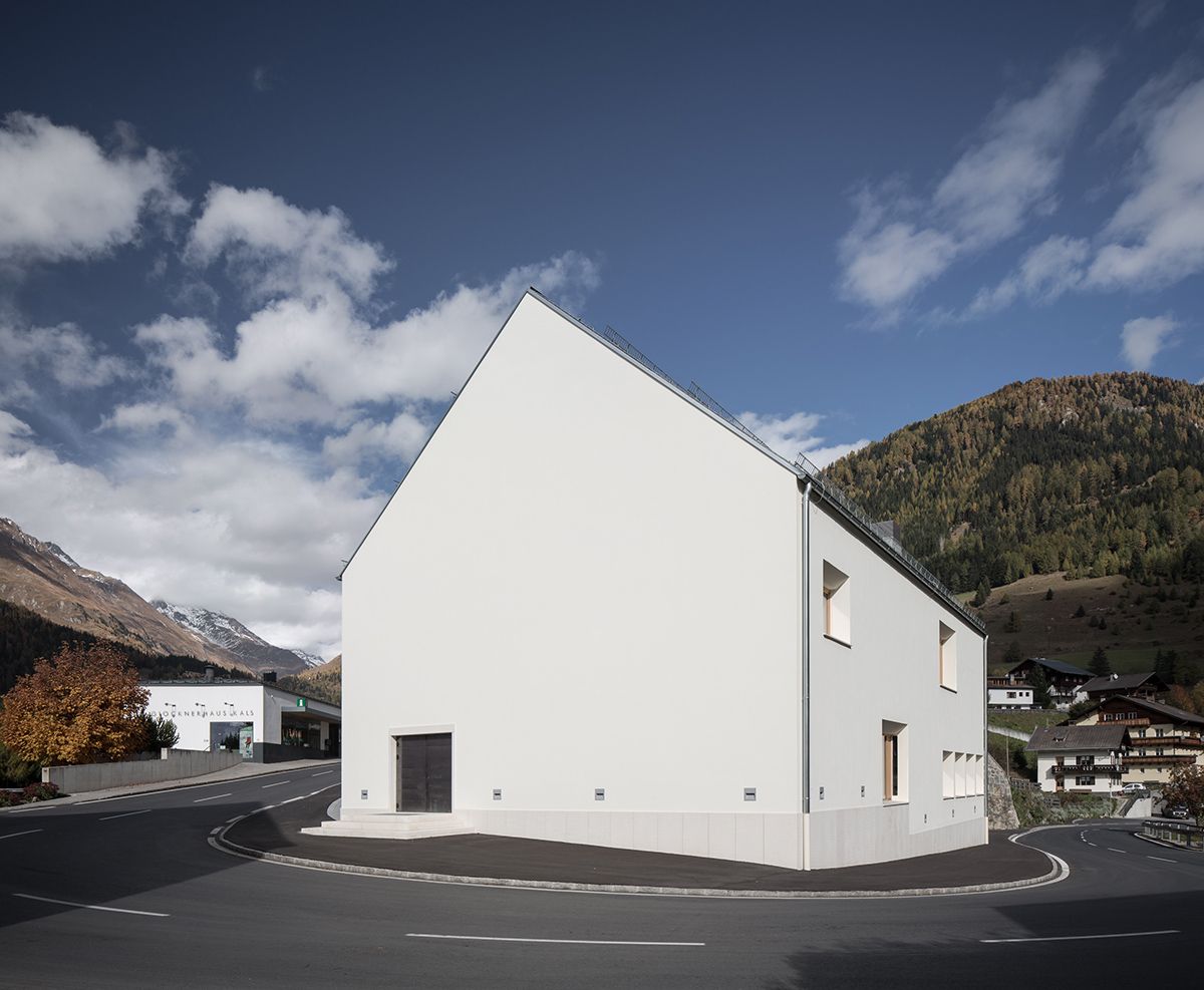 Modernes weißes Gebäude an einer kurvigen Straße, umgeben von Bergen und Herbstbäumen unter einem teilweise bewölkten Himmel.