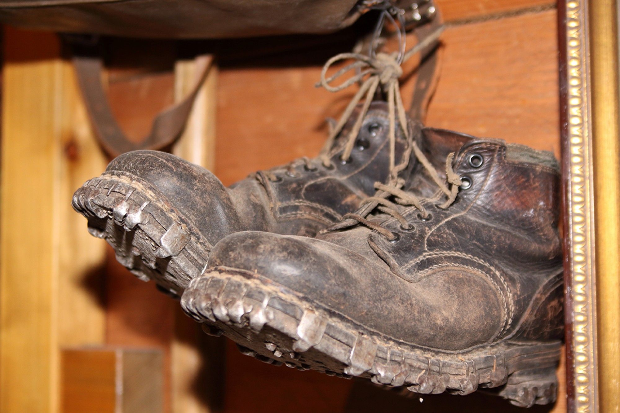 An einer Holzwand hängen abgenutzte braune Lederstiefel mit rauen Sohlen und ausgefransten Schnürsenkeln, die auf eine abenteuerliche Geschichte hinweisen.