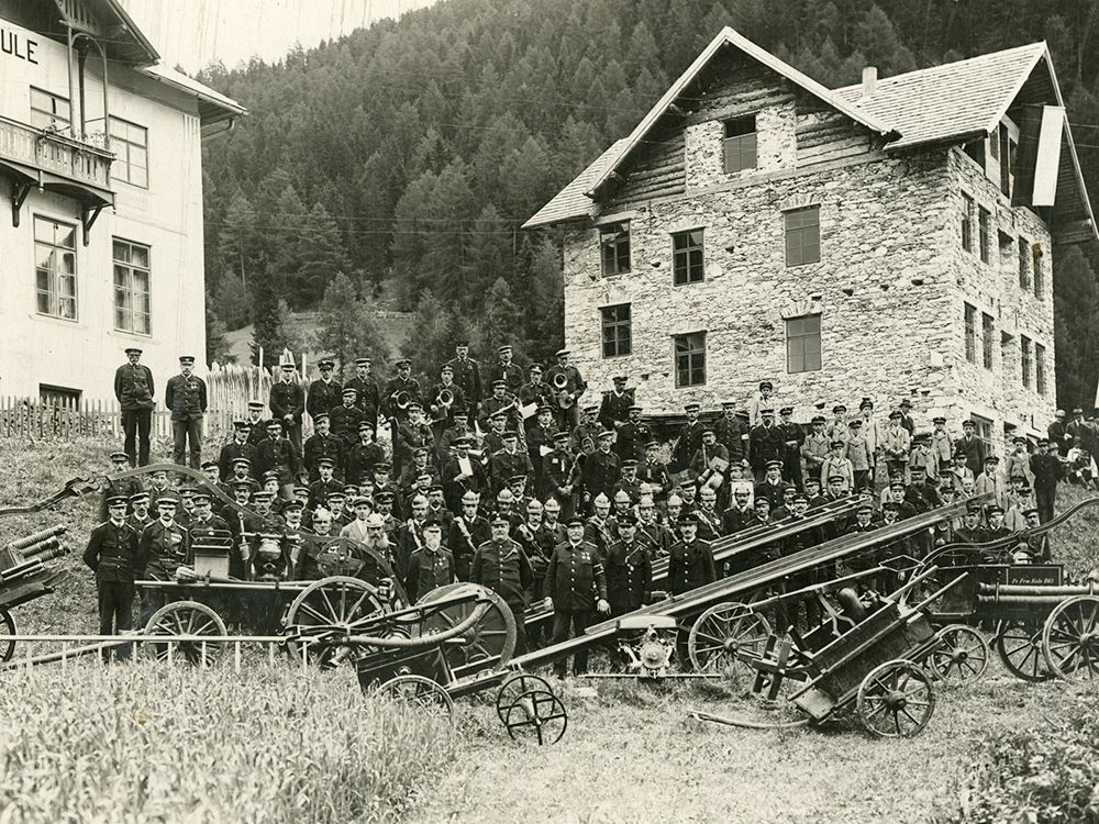 Eine große Gruppe von Uniformierten posiert im Freien in der Nähe eines steinernen Gebäudes, vor dem alte Feuerwehrgeräte ausgestellt sind.