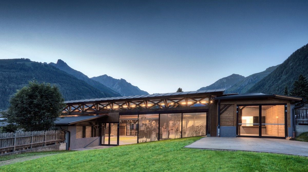 Modernes Holzgebäude mit großen Glasfenstern, vor der Kulisse majestätischer Berge in der Dämmerung, umgeben von grünem Gras.